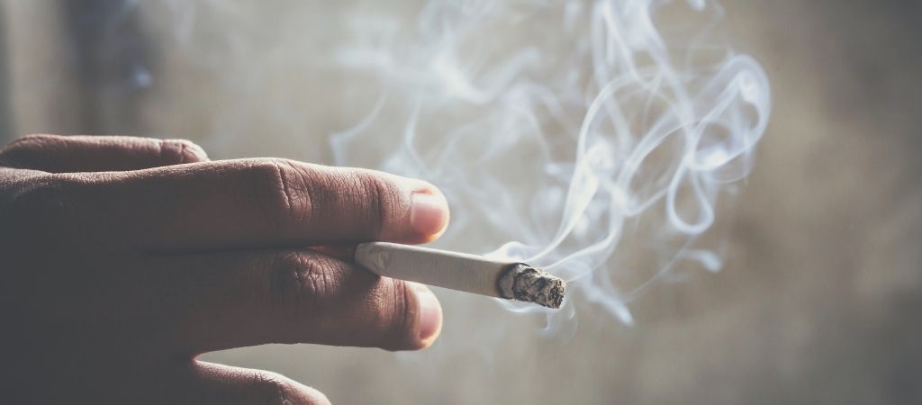 Mano sosteniendo un cigarrillo encendido con humo, simbolizando cómo afecta el tabaco a tus encías y dientes
